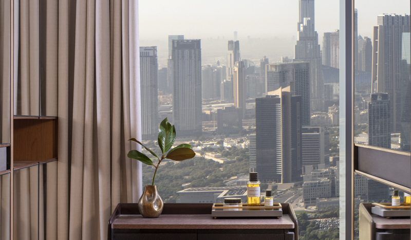 Un bureau moderne dans un appartement de Dubaï comprenant un bureau en bois et un petit tabouret, élégamment placés près d'une grande fenêtre offrant une vue panoramique sur les toits de la ville avec des gratte-ciel, dont un imposant