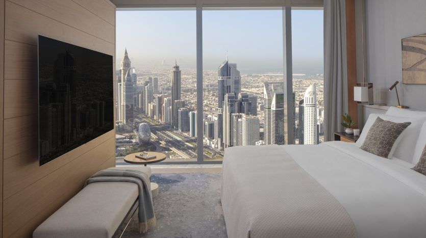 Une chambre d'hôtel luxueuse comprenant un grand lit avec une tête de lit moelleuse, une literie d'un blanc éclatant et un panneau affichant un paysage urbain. Une télévision murale se trouve en face du lit et de grandes fenêtres offrent un