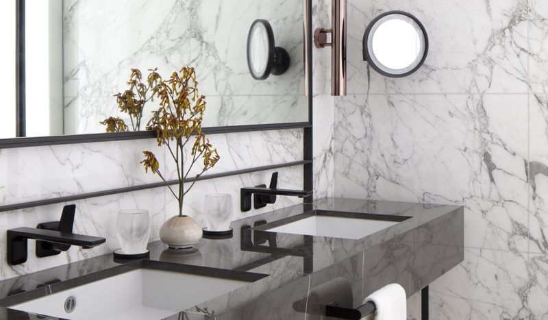 Une salle de bains luxueuse dans un appartement de Dubaï, avec des murs et un plan de vasque recouverts de marbre blanc, comprenant une douche aux parois de verre, un grand miroir et d'élégants lavabos. Des serviettes aux tons neutres complètent le