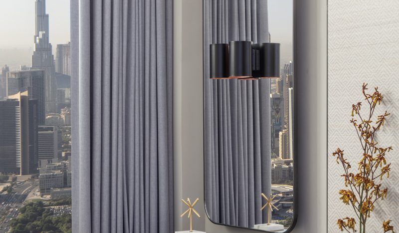 Un coin de chambre moderne avec une fenêtre offrant une vue sur Burj Khalifa, des rideaux gris, un petit bureau avec des objets décoratifs et une chaise dans un appartement de Dubaï.