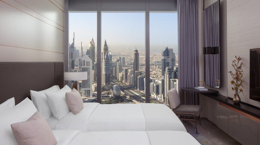 Chambre d'hôtel luxueuse avec un grand lit face à une baie vitrée offrant une vue panoramique sur les toits de la ville moderne de Dubaï pendant la journée.