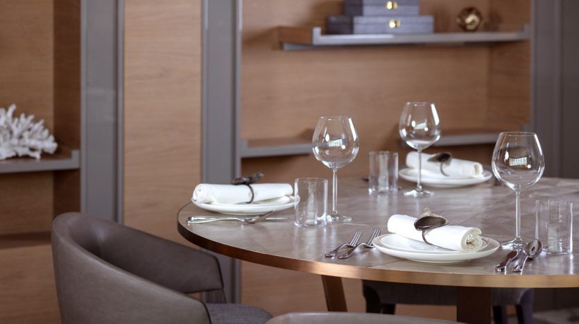 Table à manger élégante avec assiettes blanches, serviettes pliées, verres à vin et couverts dans un appartement de Dubaï doté d'un mobilier en bois et d'un éclairage tamisé.