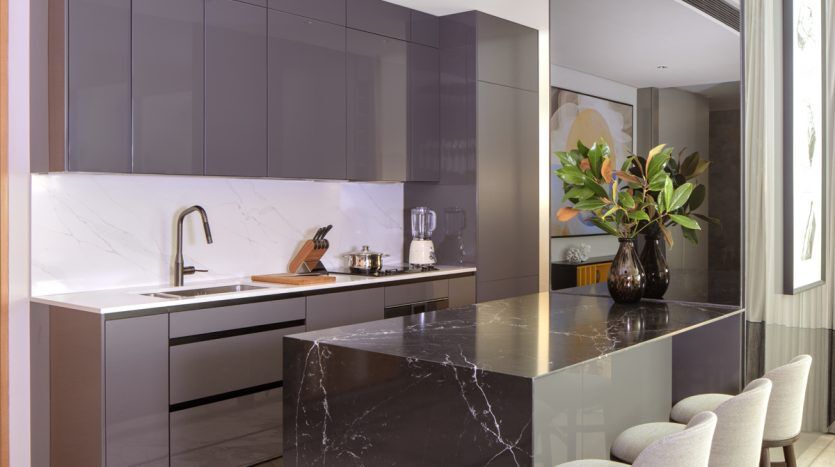 Une cuisine moderne dans une villa à Dubaï, comprenant un grand îlot en marbre, des tabourets hauts, des armoires grises sophistiquées et des appareils électroménagers en acier inoxydable, complétés par la lumière naturelle et un vase de feuilles vertes.