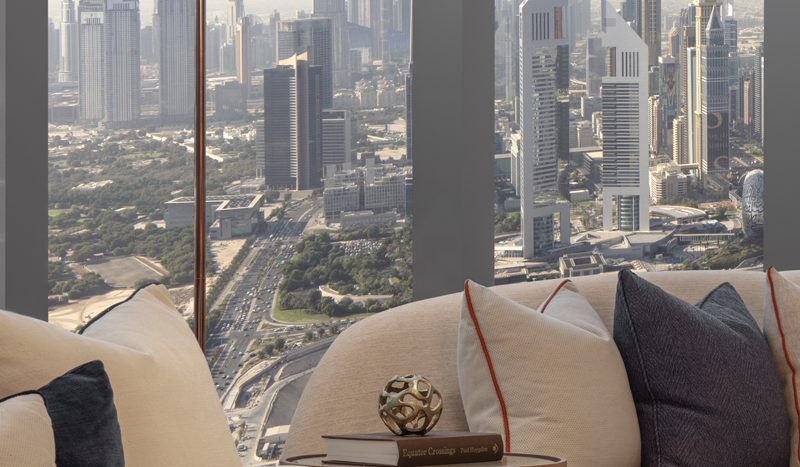 Vue depuis le salon d'un appartement de grande hauteur donnant sur les toits de la ville avec des gratte-ciels modernes et des routes de Dubaï, avec des canapés confortables et une décoration tendance au premier plan.