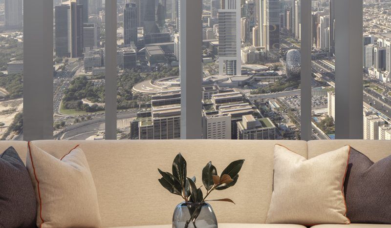 Un salon moderne avec un grand canapé et une table basse ornée d'une plante dans un vase, offrant une vue panoramique sur les toits de Dubaï à travers des baies vitrées.