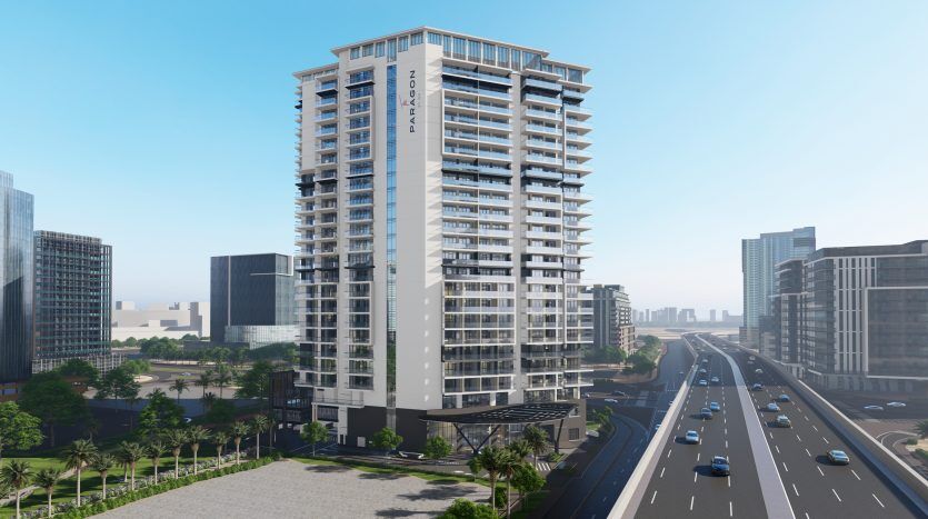 Un grand immeuble d'appartements moderne doté de nombreux balcons se trouve à Dubaï, à côté d'une autoroute où circulent des voitures, entouré d'autres bâtiments urbains sous un ciel dégagé.