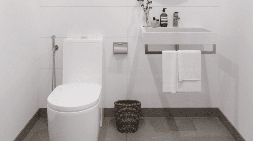 Intérieur de salle de bains moderne dans un appartement de Dubaï avec toilettes blanches, lavabo mural, miroir rond et petite plante sur une vanité. Un panier en osier est placé près de l'évier.
