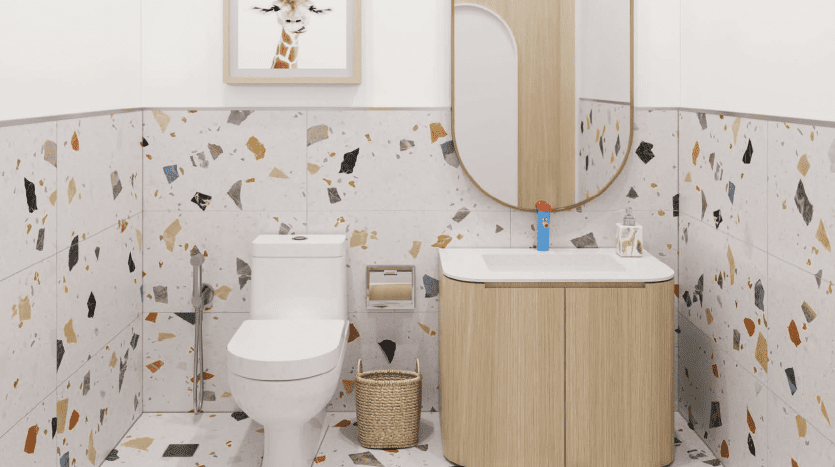 Une salle de bains moderne avec un mur carrelé en terrazzo avec des taches colorées, équipée de toilettes blanches, d'une vanité en bois surmontée d'un miroir ovale et d'un petit panier en osier. UN