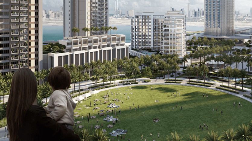 Une femme tenant un enfant en bas âge regarde un parc urbain animé entouré de grands bâtiments modernes, d'une verdure luxuriante et de nombreuses personnes profitant du plein air à Dubaï.