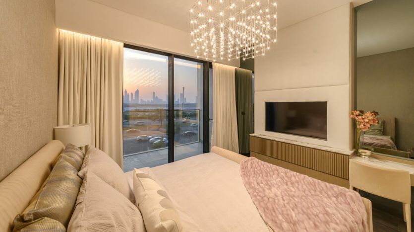 Chambre moderne avec de grandes fenêtres mettant en valeur les toits de la ville, un lit moelleux avec une couverture rose, une télévision montée et un lustre saisissant, créant une ambiance chaleureuse dans une propriété d'investissement à Dubaï.