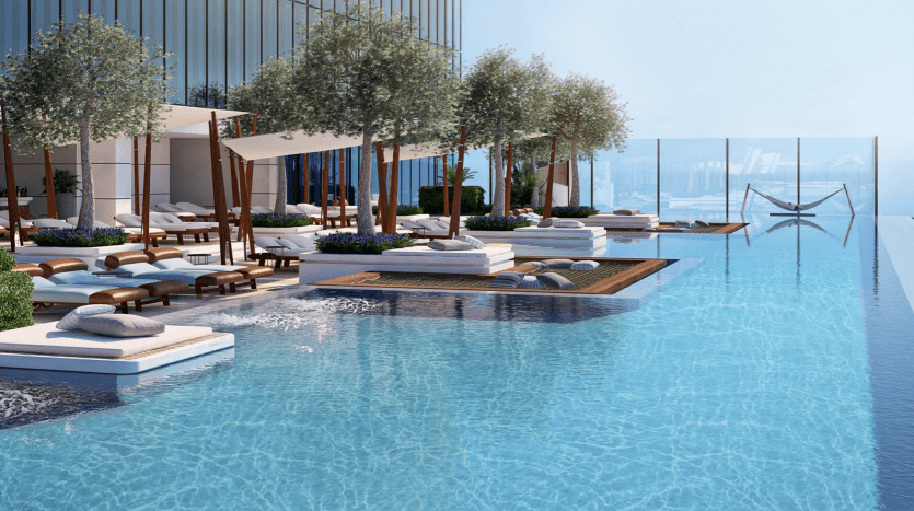 Une luxueuse piscine à débordement sur le toit avec de l'eau bleu clair, entourée d'arbres, de chaises longues et de cabanes, donnant sur une vue panoramique sur l'océan depuis un emplacement prestigieux de l'immobilier de Dubaï.
