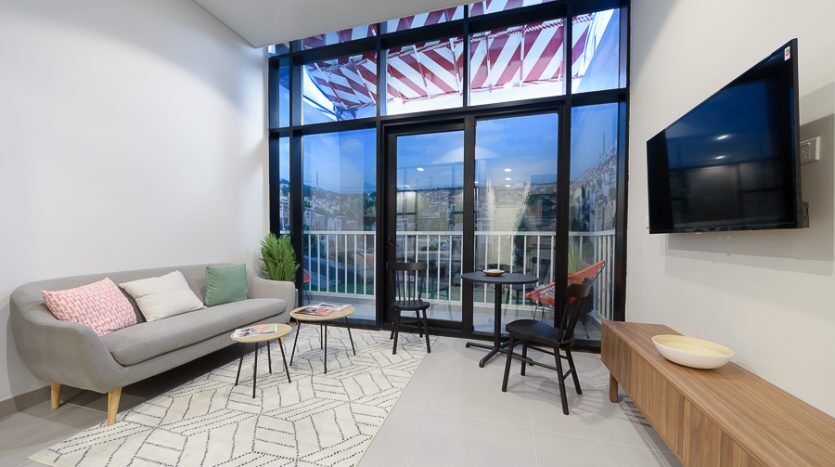 Salon moderne avec un canapé gris, un tapis à motifs et des meubles en bois, doté de grandes fenêtres avec vue sur un drapeau américain et d'une télévision à écran plat. Idéal pour investissement Dubaï.