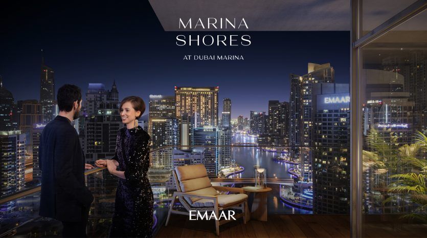 Un homme et une femme se tiennent sur un balcon offrant une vue nocturne sur la marina de Dubaï. L'horizon de la ville est illuminé et le texte superposé annonce "Marina Shores at Dubai Marina by Emaar - an