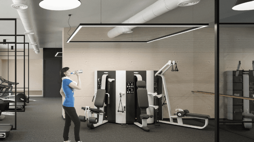 Une femme vêtue d'une chemise bleue et d'un pantalon noir buvant de l'eau dans une salle de sport moderne avec divers appareils d'exercice et de grands miroirs sur les murs à Dubaï.