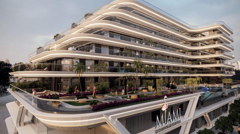 Un bâtiment moderne de plusieurs étages en bord de mer avec de vastes balcons et terrasses incurvés, entouré d'une verdure luxuriante et de personnes profitant des espaces extérieurs. Le panneau indique « Résidences de Dubaï.