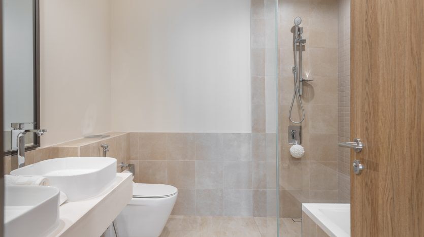 Une salle de bains moderne comprenant un lavabo blanc, des toilettes et une douche à l'italienne avec portes vitrées, le tout sur des murs et un sol carrelés beiges. Les armoires en bois naturel ajoutent de la chaleur à la villa à Dubaï.