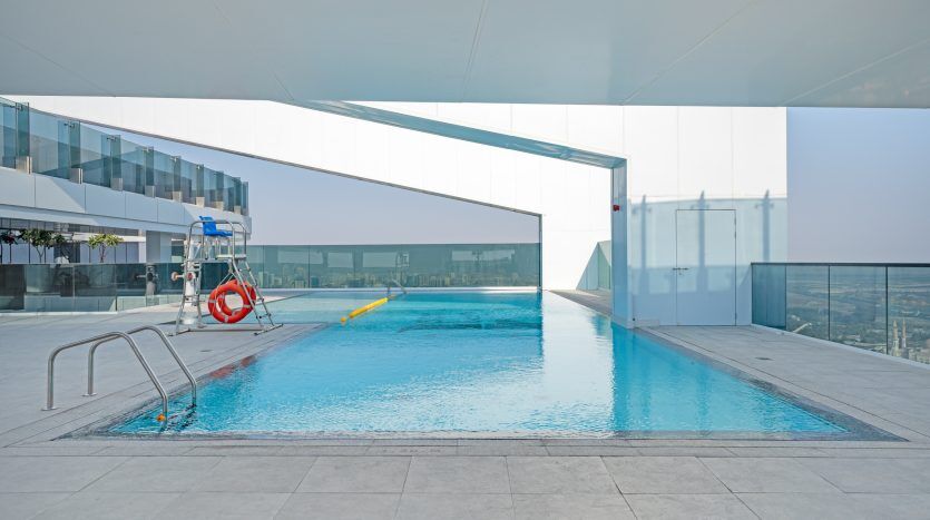 Une piscine moderne sur le toit d'une villa de Dubaï au design élégant et minimaliste, avec une eau bleu clair, entourée d'une terrasse blanche et de barrières de verre, sous un ciel clair et lumineux.
