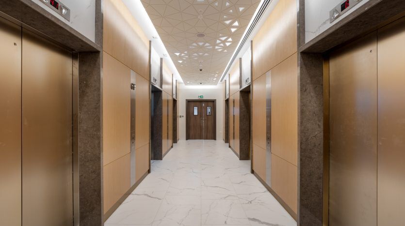 Un hall d'ascenseur moderne dans une villa de Dubaï comprenant quatre ascenseurs aux finitions en bois et métalliques, des sols en marbre blanc et un plafond à motifs géométriques. La lumière est répartie uniformément le long du couloir.