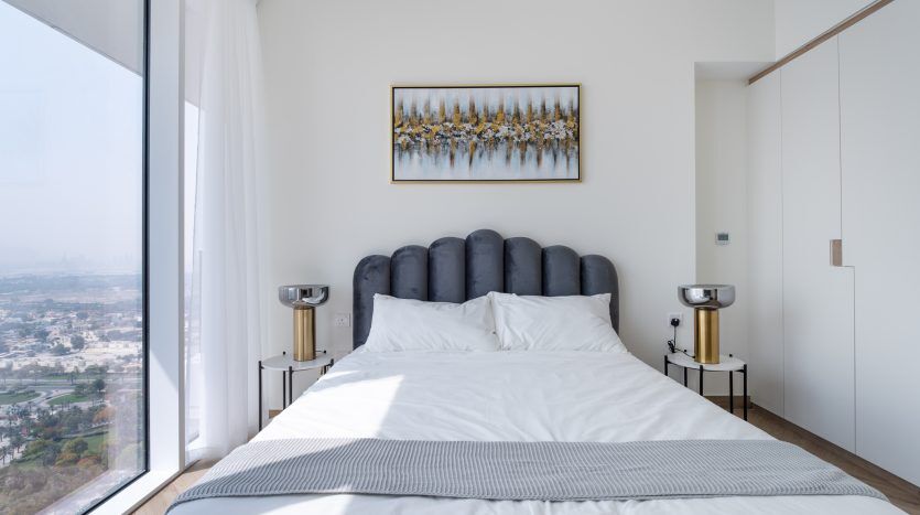 Une chambre moderne dans une villa de Dubaï avec un grand lit, une tête de lit capitonnée et une vue panoramique sur la ville à travers des baies vitrées. Des œuvres d'art sont suspendues au-dessus du lit et de deux tables de nuit sur le flanc