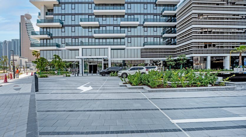 Un paysage urbain moderne à Dubaï avec un trottoir à motifs géométriques, des jardinières vertes luxuriantes et des bâtiments élégants avec des balcons horizontaux. Une voiture de luxe est visible près de l'entrée de l'un