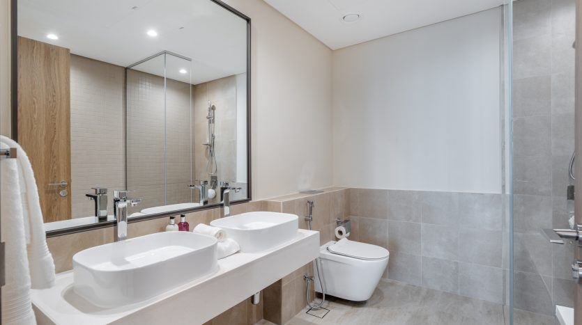 Intérieur de salle de bains moderne dans un appartement de Dubaï comprenant deux lavabos, un grand miroir et des toilettes. Tons neutres avec murs carrelés et détails d'armoires en bois.