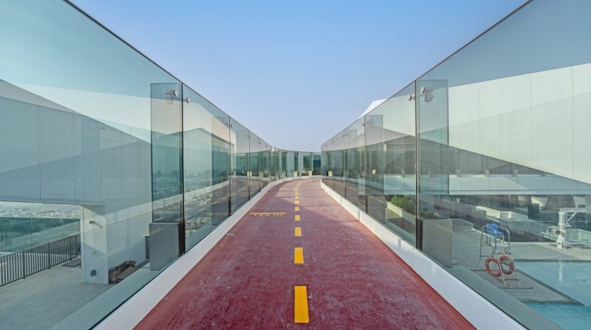Un pont piétonnier moderne avec un sol rouge et des lignes pointillées jaunes, entouré de barrières de verre transparentes, s'étendant vers un ciel bleu à Dubaï.