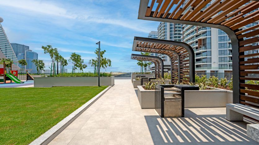 Un parc urbain moderne avec des pergolas projetant des ombres géométriques sur une allée pavée, entouré d'une verdure luxuriante et d'immeubles de grande hauteur contemporains, surplombant un front de mer lointain à Dubaï.