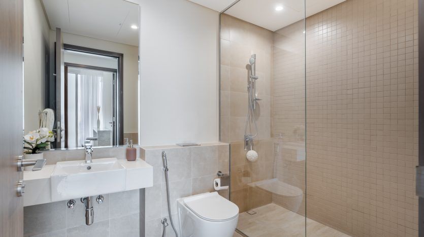 Intérieur de salle de bains de villa moderne à Dubaï avec double vasque, grand miroir, toilettes et douche à l'italienne entourée de portes en verre. Conception de carrelage neutre avec lumière naturelle.