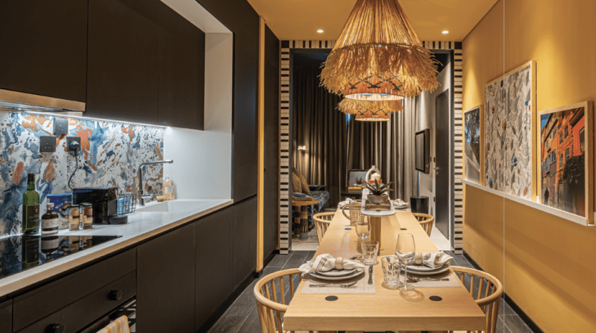 Salle à manger moderne comprenant une longue table en bois pour quatre personnes, flanquée de chaises élégantes sous un lustre unique, avec des œuvres d'art vibrantes sur les murs et un éclairage jaune confortable dans une luxueuse villa de Dubaï.