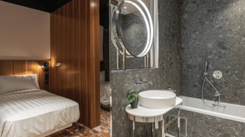 Une villa moderne à Dubaï comprenant un lit confortable avec une tête de lit en bois à gauche et une élégante salle de bains avec un lavabo blanc, un miroir rond et une baignoire à droite.