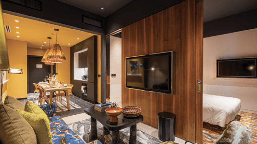 Intérieur de suite d'hôtel moderne comprenant un salon confortable avec un canapé vert et un tapis bleu unique, ainsi qu'une chambre séparée visible à travers une cloison en bois avec un lit blanc. Décoration lumineuse et élégante, parfaite pour un