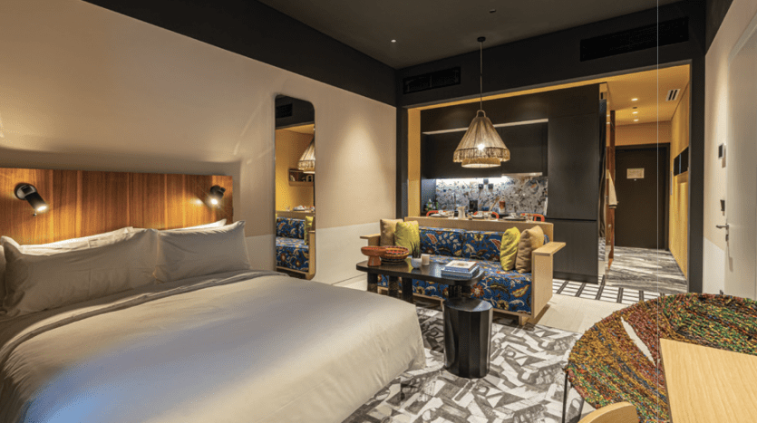 Chambre d'hôtel moderne dotée d'un grand lit avec une literie blanche, d'un coin salon coloré avec des chaises à motifs et d'un éclairage décoratif. La villa intègre des accents de bois et un sol élégant.