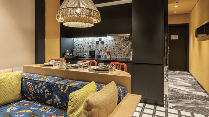Un coin repas élégant dans un appartement de Dubaï avec une banquette à motifs bleus et jaunes, un lustre élégant, un sol monochrome et une fresque murale vibrante représentant des scènes naturelles.