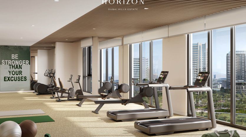 Une salle de sport moderne dotée de grandes fenêtres donnant sur un paysage urbain de Dubaï, comprenant des tapis roulants, des rameurs, des tapis de yoga et des ballons d'exercice. Le mur a une citation inspirante.