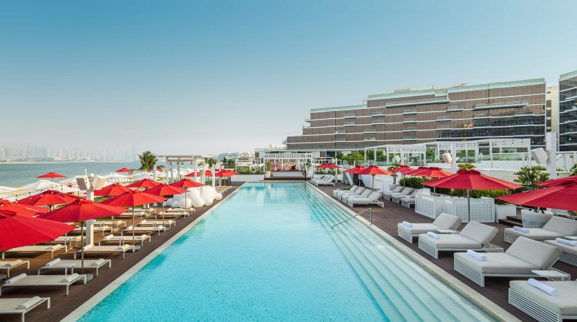 Piscine extérieure luxueuse dans un complexe, flanquée de chaises longues blanches et de parasols rouges, donnant sur des bâtiments modernes avec un horizon urbain à Dubaï.