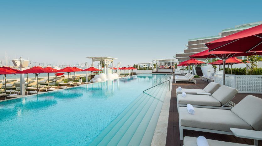 Une luxueuse piscine extérieure aux eaux bleu clair, bordée de chaises longues blanches et de parasols rouges sous un ciel lumineux. Le cadre comprend des bâtiments blancs modernes et des allusions à une marina au loin,