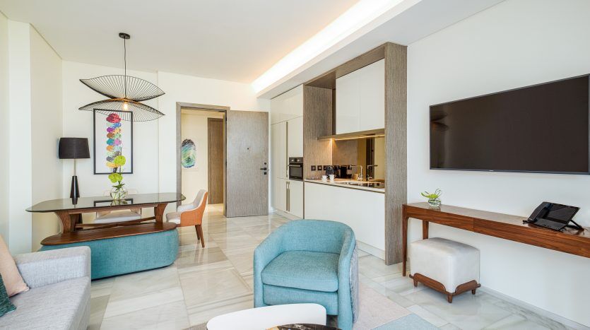 Salon moderne dans un appartement de Dubaï avec un mobilier élégant comprenant un canapé turquoise, des chaises blanches, une table en bois et une télévision murale. Cuisine ouverte visible en arrière-plan. Ambiance lumineuse et aérée.