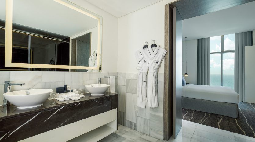 Une salle de bains luxueuse d'hôtel comprenant deux vasques blanches sur un comptoir en marbre, un grand miroir, des peignoirs accrochés à la porte, menant à une chambre de style appartement avec vue sur l'océan à Dubaï.