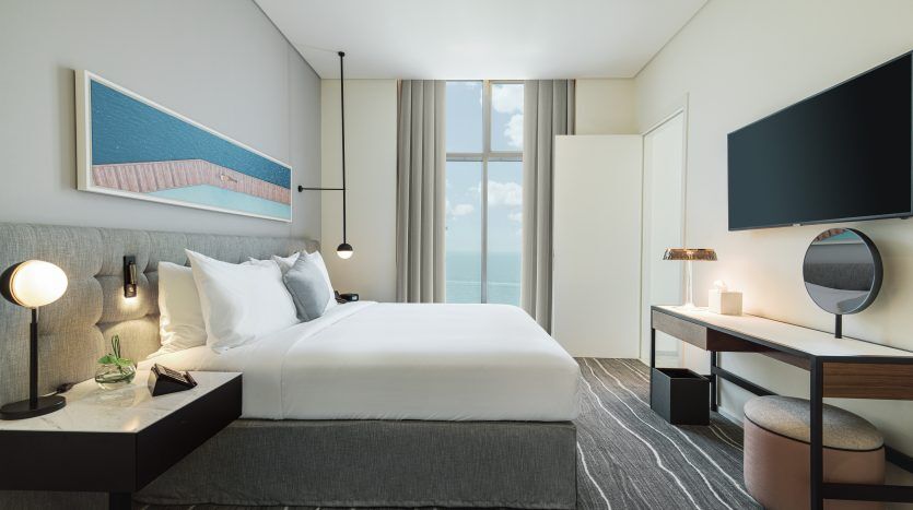 Chambre d'hôtel moderne comprenant un lit simple bien fait, un bureau avec un miroir rond, une grande fenêtre avec vue sur l'océan, un tapis rayé et une décoration minimaliste dans un appartement de Dubaï.