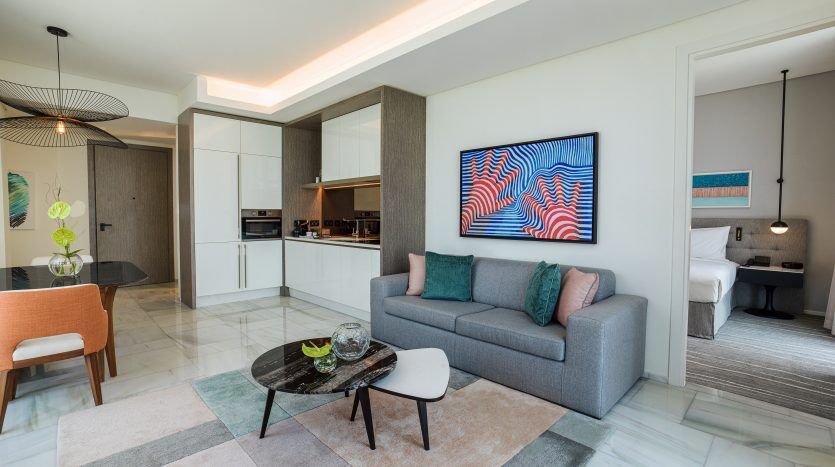 Salon moderne relié de manière transparente à une cuisine et un aperçu de la chambre dans un appartement à Dubaï. Comprend un canapé gris, des œuvres d'art colorées, un mobilier minimaliste et un éclairage élégant.