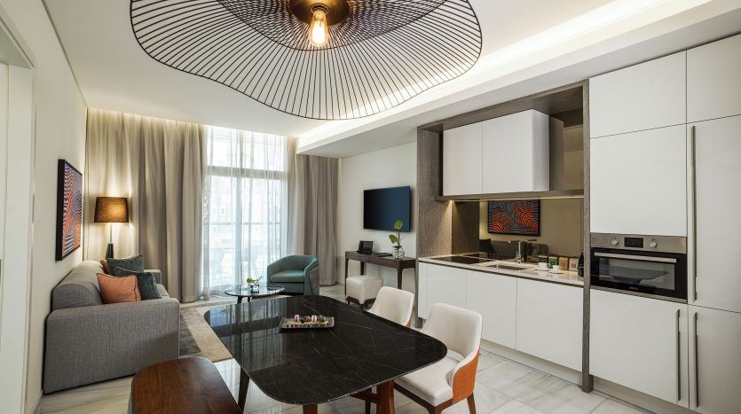 Un appartement moderne à Dubaï comprend une cuisine élégante, une table à manger ovale avec des chaises blanches, un salon confortable avec un canapé gris et un plafonnier artistique, le tout éclairé par la lumière naturelle d'un grand