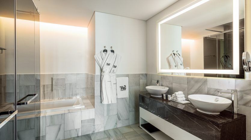 Une salle de bains luxueuse dans un appartement de Dubaï comprenant un grand miroir avec rétroéclairage LED, deux vasques blanches sur une vasque sombre et une baignoire carrelée en marbre avec des peignoirs blancs adjacents accrochés au