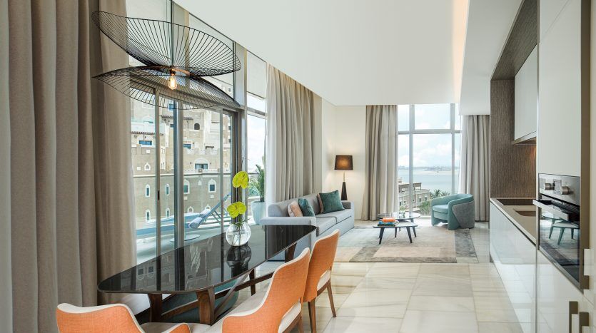 Salon moderne dans un appartement de Dubaï avec de grandes fenêtres offrant une vue sur la ville, comprenant un canapé sectionnel, une table basse en verre, un coin repas avec des chaises orange et un ventilateur de plafond décoratif. Ambiance lumineuse et aérée.