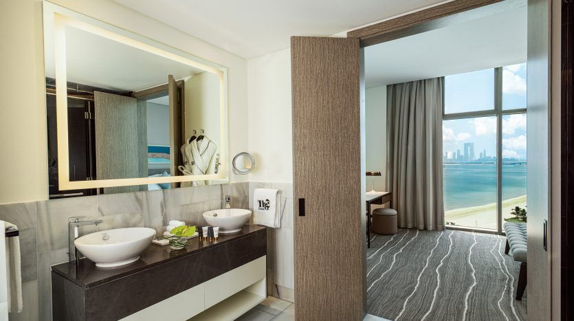Salle de bains d'hôtel moderne avec double vasque, grand miroir et vue sur la ville à travers la fenêtre d'un appartement adjacent à Dubaï. Des couleurs neutres et des éléments de design élégants créent une atmosphère luxueuse.