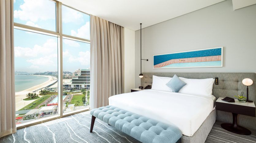 Une chambre d'hôtel moderne avec une grande fenêtre offrant une vue panoramique sur une plage de sable fin et une eau bleue. La chambre dispose d'un lit moelleux, d'une décoration élégante et d'une œuvre d'art du littoral de Dubaï au-dessus du lit.