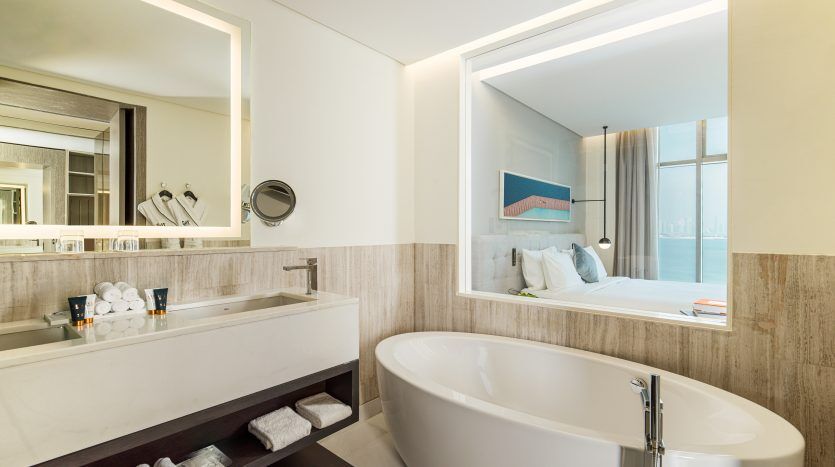 Une salle de bains moderne dans une villa de Dubaï, dotée d'une baignoire îlot, d'un grand miroir et de comptoirs blancs, avec un aperçu de la vue sur l'océan à travers la fenêtre d'une chambre.