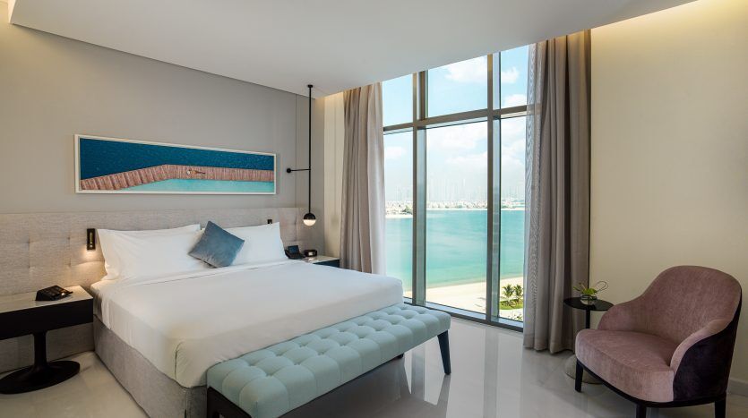Une villa moderne et lumineuse à Dubaï comprenant un grand lit aux accents bleus, un fauteuil violet et une baie vitrée donnant sur une vue ensoleillée sur la mer.