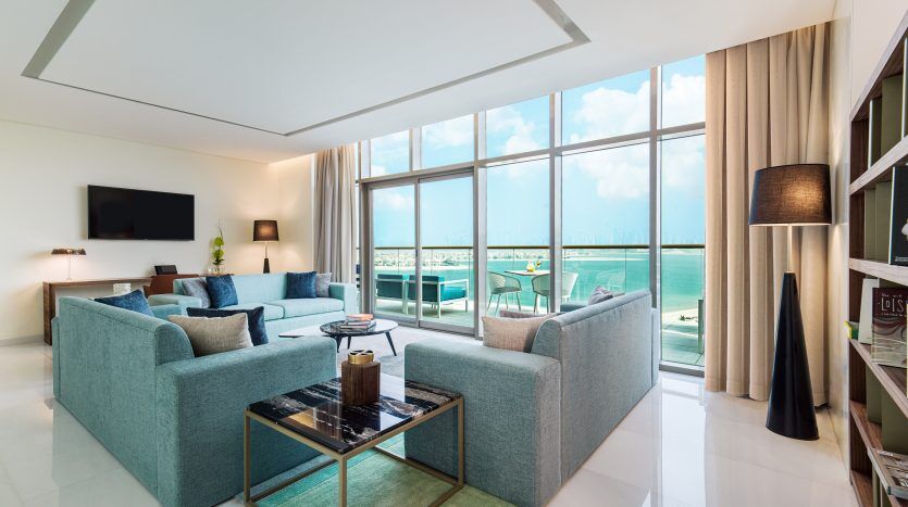Salon moderne avec de grandes fenêtres donnant sur l'océan, comprenant deux canapés bleus, une table basse en verre et une étagère. Espace lumineux et aéré avec murs blancs et design élégant dans un appartement Dubaï