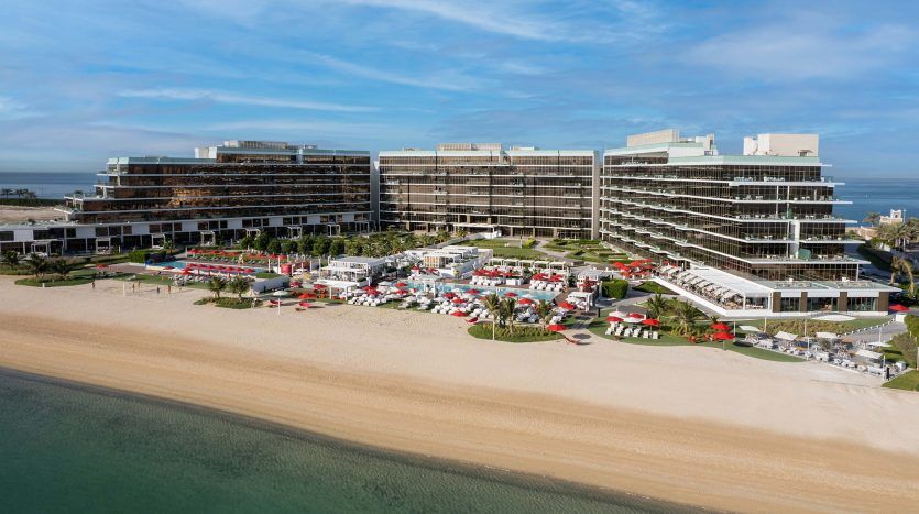 Vue aérienne d'un complexe hôtelier moderne à plusieurs niveaux en bord de mer avec parasols rouges et chaises longues sur une plage de sable adjacente aux eaux bleues calmes, idéale pour un investissement à Dubaï.