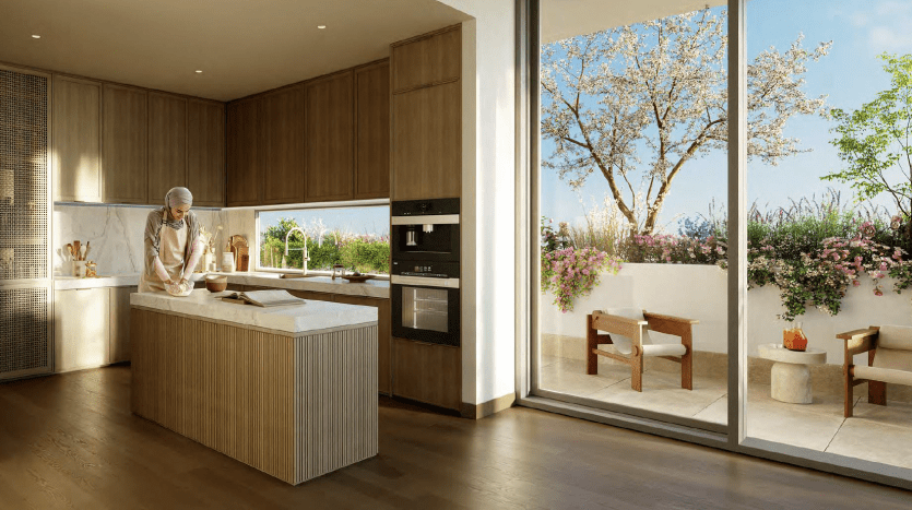 Une cuisine moderne dans une villa de Dubaï avec une personne qui cuisine au comptoir. La pièce dispose d'armoires en bois, d'appareils électroménagers intégrés et de grandes fenêtres donnant sur un patio avec des arbustes à fleurs et des sièges.
